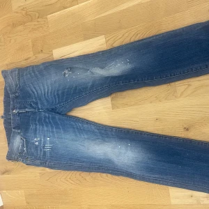 Blå jeans från Dsquared2, storlek 46 - Snygga blå jeans från Dsquared2 med slitningar och färgstänk för en cool, avslappnad vibe. Klassisk femficksmodell med broderad logga på bakfickan. Jeansen har rak passform och är tillverkade i bomull. Perfekta för dig som gillar streetstyle.