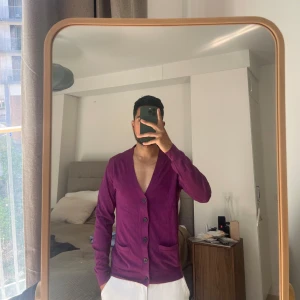 Lila kofta från H&M - Säljer en stilren lila kofta från H&M i silk bomullsmaterial. Koftan har en v-ringad design med knappar framtill och långa ärmar. Perfekt för lager-på-lager under kyligare dagar.