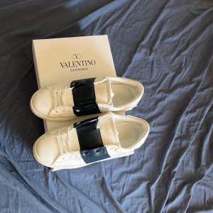 Valentino Garavani vita sneakers - Snygga vita sneakers från Valentino Garavani med bred svart rem över vristen och klassisk snörning. Skorna har en stilren och modern design i skinn som passar perfekt till streetwear eller en clean look. Pris kan diskuteras. Storlek 42 men kan även passa 43 bilderna blev jätte suddiga har inte den bästa kameran!
