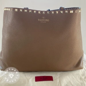 Valentino Garavani Rockstud Tote Bag axelväska - Säljer en snygg brun axelväska från Valentino Garavani med ikoniska guldfärgade nitar längs kanten. Väskan är tillverkad i exklusivt skinn med en mjuk, kornig struktur och har magnetlås. Insidan är fodrad i satin och har innerficka med dragkedja. 