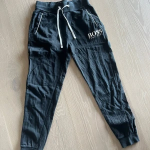 Svarta mjukisbyxor från Hugo Boss - Svarta mjukisbyxor från Hugo Boss med vit logga på låret och vita snören i midjan. Byxorna har ribbade muddar vid bensluten och vita detaljer vid fickorna. Storlek M, men små i storleken. 