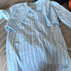 Ljusblå randig skjorta med knappar - Snygg ljusblå skjorta med vita ränder och knappar framtill. Skjortan har korta ärmar och en avslappnad passform, perfekt för varmare dagar. Tillverkad i ett tunt och svalt material som känns skönt mot huden.