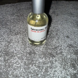 Sørensen 26.0 Extrait de Parfum - Parfymflaska från Sørensen med doften 26.0 Extrait de Parfum. Flaskan är rund och genomskinlig med ljusgul vätska och har en stilren, silverfärgad spraykork. Etiketten är vit med svart och röd text för en minimalistisk look. Luktar vädligt mycket vanilj. Har testat och spruta med den 1 gång 