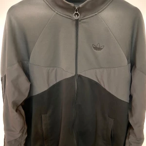 Grå track jacket från Adidas - Snygg grå track jacket från Adidas med diskret logga på bröstet och hel dragkedja framtill. Jackan har hög krage, två fickor och coolt färgblocksmönster i olika grå nyanser. Perfekt för en sportig och avslappnad stil.