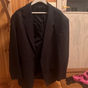 Svart randig kavaj med knappar - Klassisk svart kavaj med diskreta tunna ränder och två knappar framtill. Kavajen har breda slag, innerfoder och fickor med lock. Perfekt för en stilren look och passar till skjorta eller t-shirt. Använd fåtal gånger