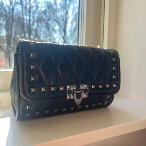 Svart axelväska med nitar - Snygg svart axelväska i quiltat mönster med silverfärgade nitar och kedja. Väskan har en stilren design med ett lås framtill. Man kan både göra axelbandet långt och kort. 