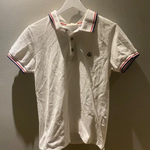 Vit kortärmad pikétröja från Moncler - Säljer en stilren vit pikétröja från Moncler med korta ärmar och klassisk krage. Tröjan har röd och blå randdetalj på krage och ärmslut samt knappar framtill. Perfekt för en avslappnad men ändå snygg look. Lite smutsig 