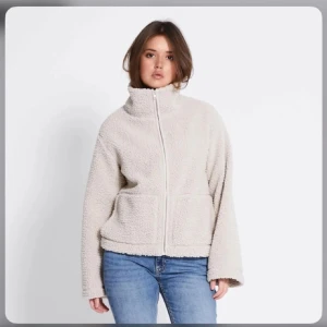 Beige teddyjacka med dragkedja - Mysig beige teddyjacka med dragkedja och hög krage. Perfekt för kyliga dagar med sin fluffiga och varma design. Jackan har en avslappnad passform och två praktiska fickor framtill.