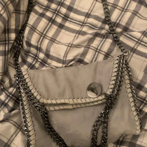 Grå axelväska med kedjedetaljer - Snygg grå axelväska med kedjedetaljer och dekorativa sömmar. Väskan har en praktisk magnetknapp för enkel stängning och en stilren design som passar till många tillfällen. Perfekt för att ge din outfit en cool touch.