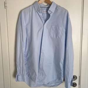 Ljusblå skjorta från Lacoste - Säljer en stilren ljusblå skjorta från Lacoste med klassisk krage och knappstängning. Skjortan har en bröstficka med det ikoniska Lacoste-emblemet broderat ovanför. Perfekt för en avslappnad men ändå elegant look. Storlek 41 vilket motsvarar L. Ligger ute en likadan i vit färg, köps båda samtidigt får du de för 700 totalt