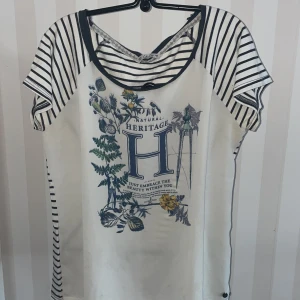 Vit och randig topp från Cotton Club - Säljer en vit topp med randiga detaljer och ett stort tryck framtill från Cotton Club. Toppen har korta ärmar och ett unikt blommönster med text. Perfekt för en avslappnad stil.