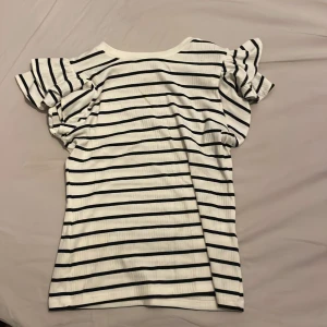 Randig topp från Holly & Whyte - Säljer en vit och svart randig topp från Holly & Whyte by Lindex. Toppen har korta ärmar med volangdetaljer och är tillverkad i ett ribbat material. 