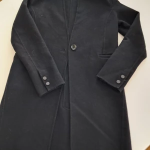 Svart kappa från Saki - Elegant svart ull kappa från Saki med enkel knäppning framtill och stilrena detaljer. Kappan har långa ärmar och en klassisk krage, perfekt för kyligare dagar. Materialet är 50% ull.