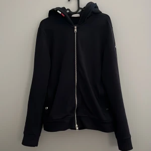 Mörkblå hoodie från Moncler - Säljer en stilren mörkblå hoodie från Moncler med dragkedja och huva. Tröjan har ett broderat Moncler-märke på ärmen och är perfekt för en casual look. Den är tillverkad i ett mjukt material som ger en bekväm passform. DM för mer bilder eller info✉️