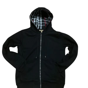 Svart hoodie från Burberry - Säljer en stilren svart hoodie från Burberry med klassiskt rutigt mönster i huvan. Tröjan har dragkedja framtill och långa ärmar. Perfekt för en avslappnad och trendig look.