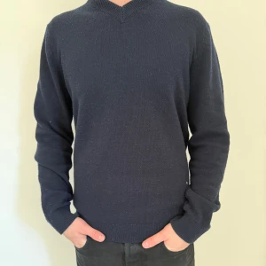 Mörkblå stickad tröja - Säljer en stilren mörkblå stickad tröja med v-ringad hals. Tröjan har långa ärmar och ribbade muddar vid ärmslut och nederkant. Perfekt för kyligare dagar.