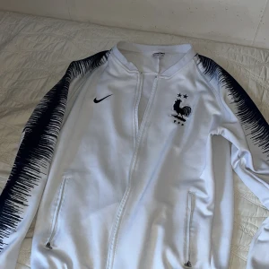 Vit träningsjacka från Nike - Säljer en vit träningsjacka från Nike med svarta detaljer och dragkedja. Jackan har ett tryck av en tupp med stjärnor på bröstet och svarta mönster längs ärmarna. Perfekt för sportiga tillfällen!
