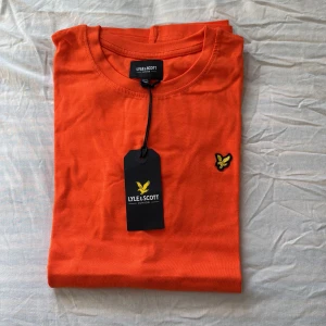 Orange t-shirt från Lyle & Scott - Snygg orange t-shirt från Lyle & Scott med deras ikoniska logga broderad på bröstet. Perfekt för en casual look. Gjord i mjukt material för extra komfort. Passar perfekt till jeans eller shorts!