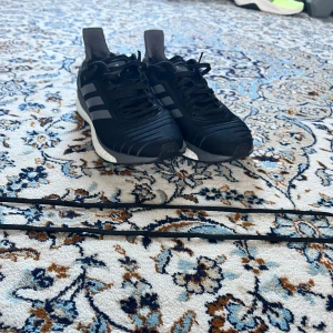 Svarta Adidas sneakers - Snygga svarta Adidas sneakers med vit Boost-sula och klassiska tre ränder på sidan. Skorna har snörning och en modern design som passar perfekt för både träning och vardag. De är bekväma och ger bra stöd för fötterna skosnörena är som ni ser klippta, men man får självklart med ett par nya snören, skorna är inköpta för 1600 kr. Men jag säljer dem för bara 250 kr. Skicka är tio av tio bara använt inomhus cirka tio gånger.