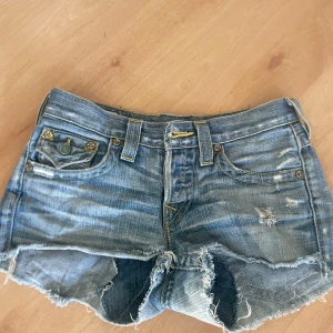 Lågmidjade blå jeansshorts  - Säljer ett par coola blå jeansshorts från True Religion med slitna detaljer och fransar. Shortsen har dekorativa knappar och broderade fickor. Perfekta för en avslappnad stil! De är 36 cm i midjemått 💕