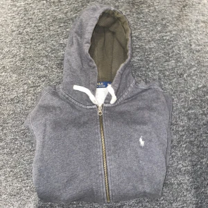 Grå hoodie från Polo Ralph Lauren - Säljer en stilren grå hoodie från Polo Ralph Lauren med dragkedja och klassisk logga på bröstet. Tröjan har långa ärmar och en praktisk huva med snören. Perfekt för en avslappnad look.