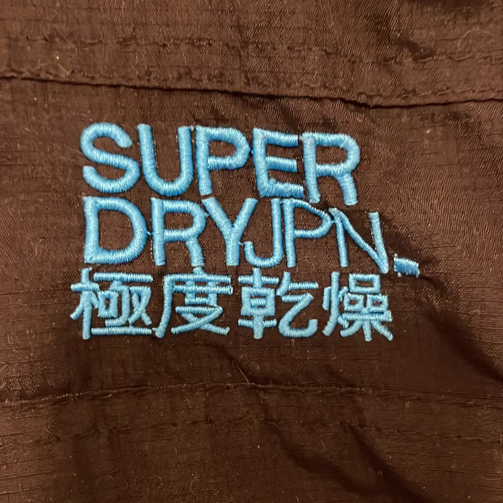 Säljer en svart vindjacka från Superdry med blå detaljer och broderad logga. Jackan har en huva och dragkedja framtill. Insidan är fodrad med blått tyg för extra komfort. Perfekt för blåsiga dagar! . Takit.