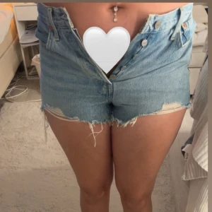 Blå jeansshorts från Levi's - Säljer ett par klassiska blå jeansshorts från Levi's. De är högmidjade med fransiga kanter och har en knappgylf. Perfekta för en avslappnad stil. Storlek 28