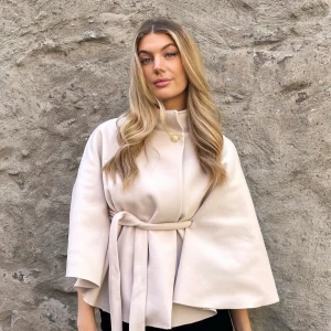 Beige kappa med bälte - Elegant beige kappa med bälte och hög krage. Jackan har en stilren design med vida ärmar och en knappdetalj vid halsen. Perfekt för en chic look. Aldrig använd, nyskick💕