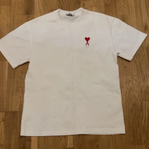 Vit t-shirt med rött hjärta - Säljer en stilren vit t-shirt med ett litet rött hjärta broderat på bröstet. Perfekt för en enkel och snygg look. T-shirten har korta ärmar och en normal passform.