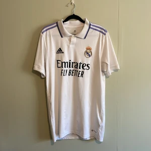  Real Madrid fotbollströja 22/23 hemma - Säljer en vit Real Madrid fotbollströja från Adidas med korta ärmar och klubbens emblem på bröstet. Tröjan har lila detaljer på axlarna och sponsorlogga framtill. Perfekt för alla fotbollsfans! ⚽