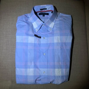 Blå rutig skjorta från Tommy Hilfiger - Ljusblå skjorta från Tommy Hilfiger. Är i nyskick bortsett från att den tråkigt nog har fått ett litet hål i ärmen. Kontakta oss gärna vid frågor