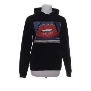 Svart hoodie med tryck från The Cool Elephant - Säljer en svart hoodie från The Cool Elephant med ett coolt tryck av röda läppar på framsidan. Tröjan har en klassisk huva med dragsko och långa ärmar. Perfekt för en avslappnad stil.