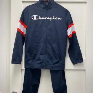 Champion, jacka + byxa för 11-12 år - Säljer ett mörkblått ”träningsset” (jacka med dragkedja och byxa med snöre) från Champion. Vita samt röda ränder på ärmarna. Jackan har en hög krage och Champion-logga tryckt på bröstet. Perfekt för sportiga aktiviteter eller en avslappnad stil. Fint skick! Storlek L för barn, 11-12 år.
