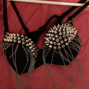 Svart BH med nitar och kedjor - Säljer en svart korsett med coola silverfärgade nitar och kedjor som hänger ner. Perfekt för en edgy look. Justerbara axelband för bästa passform.