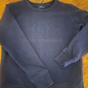 Mörkblå sweatshirt från GANT - Säljer en stilren mörkblå sweatshirt från GANT med broderad logotyp på framsidan. Tröjan har långa ärmar och ribbade muddar vid ärmslut och nederkant. .