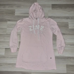 Rosa hoodie från Zink - Säljer en söt lång rosa hoodie från Zink med texten 'ZINK EST. 2015' på framsidan. Den har en stor ficka framtill och en justerbar huva med snören. Perfekt för en avslappnad stil.