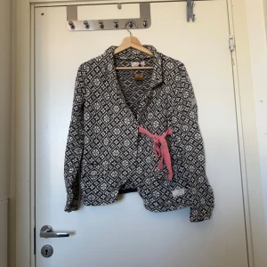 Mönstrad kofta med rosa knytband - Svartvit kofta från Odd Molly med snyggt geometriskt mönster och rosa knytband i midjan. Långärmad och mjuk, perfekt för lager på lager. Koftan har två små fickor fram och broderade rosa detaljer bak. Skön och unik stil för dig som gillar statement-plagg. Storlek 2 som enligt deras storleksguide motsvarar en storlek M.  Fint skick, men det finns små tecken på användning.  Se  bild 5.