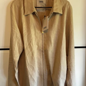 Beige overshirt i linne från OAS - Snygg beige overshirt från OAS i luftigt linne. Jackan har klassisk krage, dragkedja framtill och raka ärmar. Perfekt för lager-på-lager och chill stil. Enkel och stilren design som passar till det mesta. Ny pris ligger på 2000kr