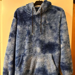Blå fluffig hoodie från Account - Supermjuk och oversized blå hoodie från Account med fluffigt teddyfleece-material. Har huva med snörning och stor magficka framtill. Perfekt för kalla dagar när du vill vara extra cozy. Snyggt mönster i olika blå nyanser.