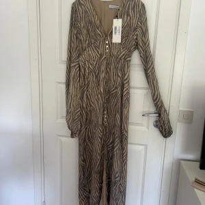 Zebramönstrad långklänning från Adoore - Helt oanvänd endast testad klänning från Adoore. Långklänning med zebramönster i beige och brunt. Klänningen har v-ringning, långa ärmar och knappar framtill. Storlek 40 men lite mindre i storlek