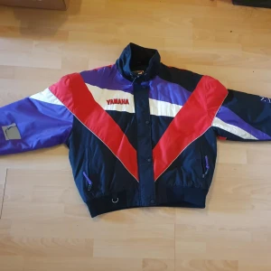 Yamaha Jacka  - Fet Yamaha vindjacka i klassisk retrostil med färgblock i rött, lila, vitt och svart. Broderad Yamaha-logga fram och stor logga bak. Jackan har hög krage, dragkedja och knappar, samt resår i midja och ärmslut. Materialet är nylon med polyesterfoder.