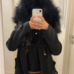 Svart parkajacka med fuskpäls från DM - Svart parkajacka från Dennis Maglic med stor huva kantad av fluffig svart fuskpäls. Jackan har snygga gulddetaljer, dragkedja framtill och dekorativa spännen. Perfekt inför höst/vinter. Köpt för 1799kr och säljs inte längre i hemsidan. 