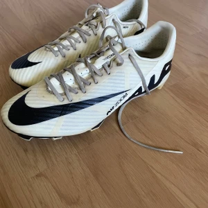 Nike Air Zoom fotbollsskor vit/svart - Säljer ett par Nike Air Zoom fotbollsskor i stilren vit färg med svarta detaljer och stor AIR-logga på sidan. Skorna har snörning, platt sula med dobbar och är tillverkade i syntetmaterial för bästa grepp på planen. Perfekta för dig som vill sticka ut på matchen.