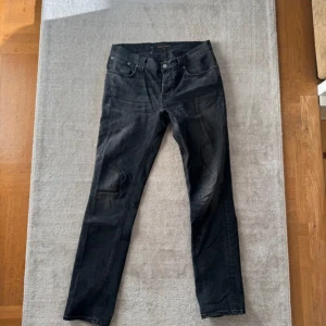 Svarta jeans från Nudie Jeans, 33/34 - Säljer ett par svarta jeans från Nudie Jeans i storlek 33/34. Jeansen har klassisk femficksdesign, raka ben och snyggt slitna detaljer. Tillverkade i mjukt denimtyg som sitter skönt och passar till allt. Perfekta för dig som gillar stilrena svarta jeans. Inga defekter. Slim fit