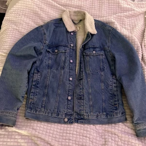 Blå jeansjacka med teddyfoder L.O.G.G. - Säljer en klassisk blå jeansjacka från L.O.G.G. med mysigt vitt teddyfoder och krage. Jackan har knappar framtill, två bröstfickor med lock och långa ärmar. Perfekt för höst och vår när du vill ha en chill och tidlös look.