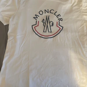 Vit Moncler t-shirt med stor logga - Vit t-shirt från Moncler med stor broderad logga i blått, rött och vitt på bröstet. Klassisk passform och rund hals. Perfekt för dig som gillar stilrena plagg med exklusiv känsla och tydlig branding.