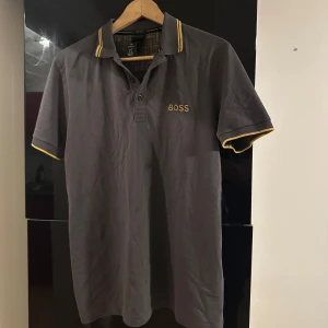 Grå BOSS pikétröja med guldiga detaljer - Snygg grå pikétröja från BOSS med guldiga detaljer på krage och ärmslut i storlek S. Tröjan är knappt använd och säljs nu pga den ej passar min stil längre. 