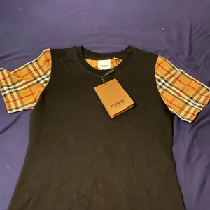 Svart Burberry t-shirt med rutiga ärmar - Svart t-shirt från Burberry med klassiska rutiga korta ärmar i beige, rött och svart. Snygg kontrast mellan den svarta kroppen och de ikoniska Burberry-rutorna på ärmarna. Rund hals och mjukt bomullsmaterial. Perfekt för dig som gillar designer och vill sticka ut.