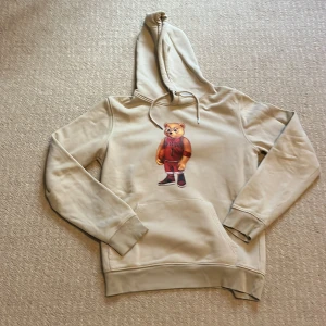 Beige hoodie Baron Filou med björntryck - Säljer en beige hoodie från Baron Filou med ett stort tryck av en basketspelande björn på bröstet. Hoodien har snörning i huvan, känguruficka och ribbade muddar. Perfekt för dig som gillar streetwear och coola prints.
