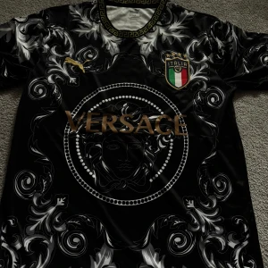 Svart Versace x Puma Italien fotbollströja - Unik svart fotbollströja med Versace-tryck och Medusa-motiv, detaljerad med guldiga inslag och Italiens landslagsemblem. Tröjan har korta ärmar, grekisk mönstrad krage och är tillverkad i ett glansigt syntetmaterial. Perfekt för dig som gillar statement-plagg och exklusiv design.